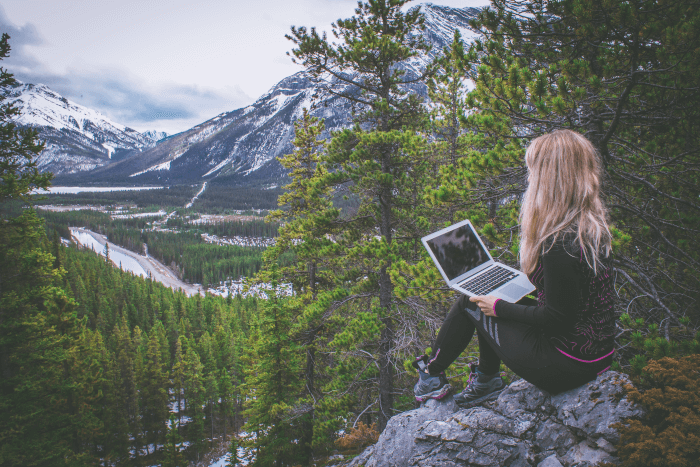 Woman in sports gear sitting on rock overlooking trees with laptop