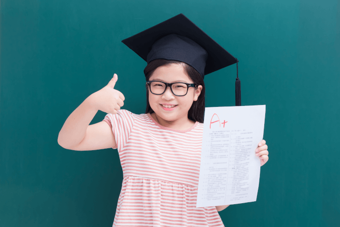 Young girl wearing graduation cap giving thumbs up and showing a+ exam paper